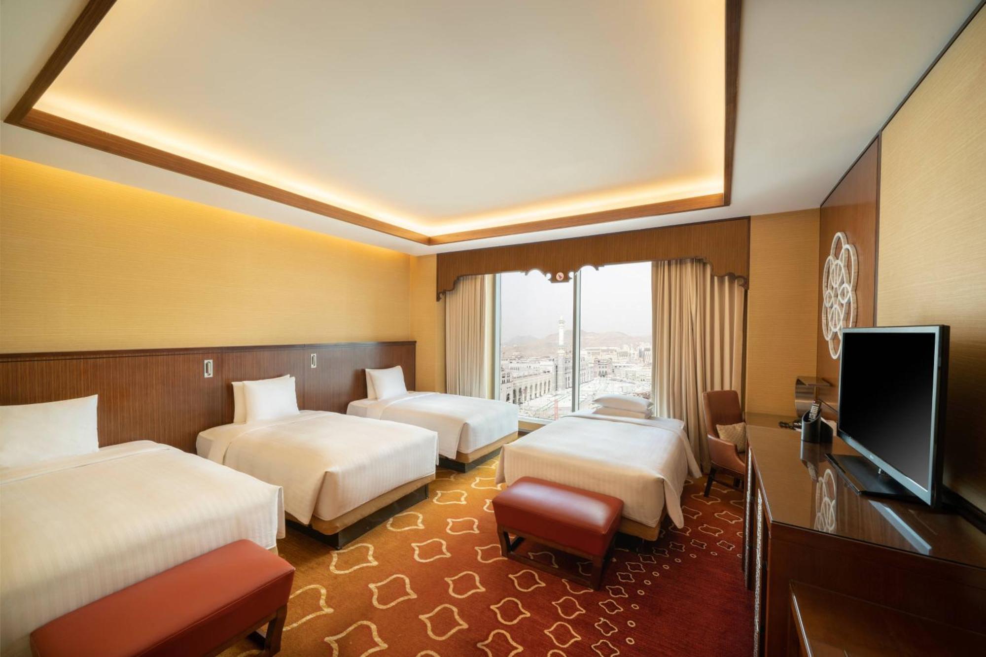Jabal Omar Marriott Hotel Makkah Mekka Buitenkant foto The photo shows a spacious hotel room featuring four beds arranged in a clean and modern style. The room has a warm color palette with yellow and brown tones. Natural light enters through large windows adorned with curtains, providing a view beyond. 
