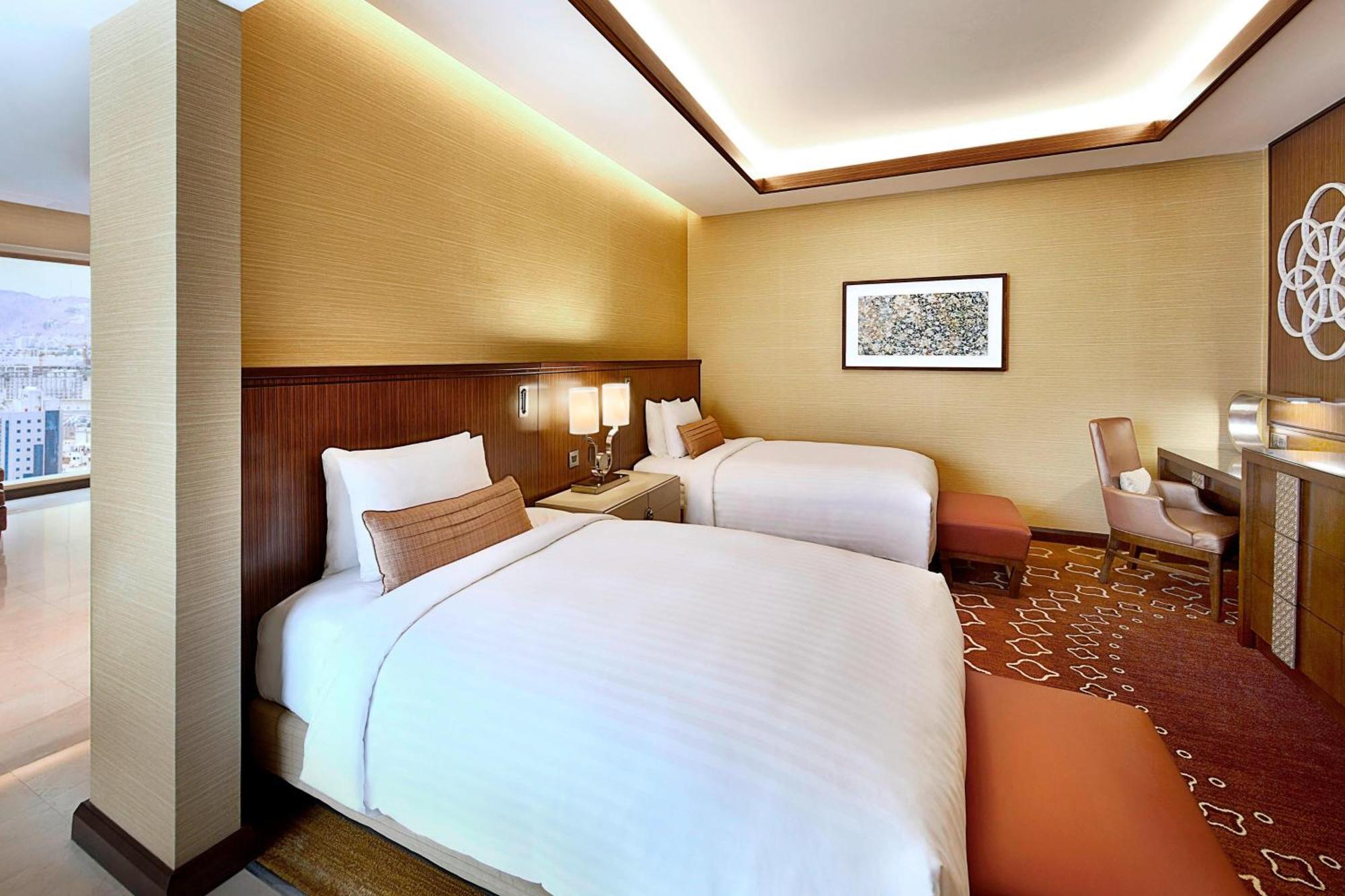 Jabal Omar Marriott Hotel Makkah Mekka Buitenkant foto The photo shows a modern hotel room featuring two beds with white linens. The room has a warm color palette, with beige walls and wooden accents. There's a piece of artwork on the wall above the beds. A small seating area is present, along with a wor